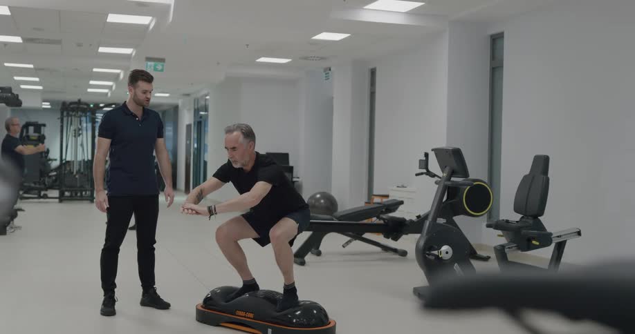 A man black shorts and a t shirt is working on his balance as he works out with a trainer in a gym