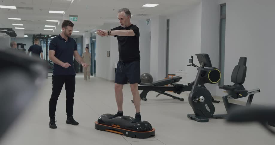 A trainer watches as a client does squats on a balancing board during therapy