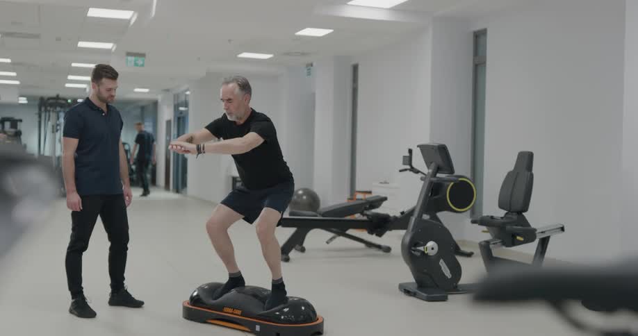Two men in a gym with one exercising with equipment and lights in the background