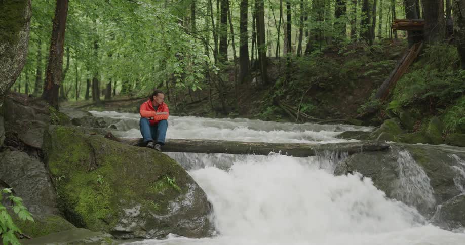 A man sits next to a river in a forest and watches the rapids