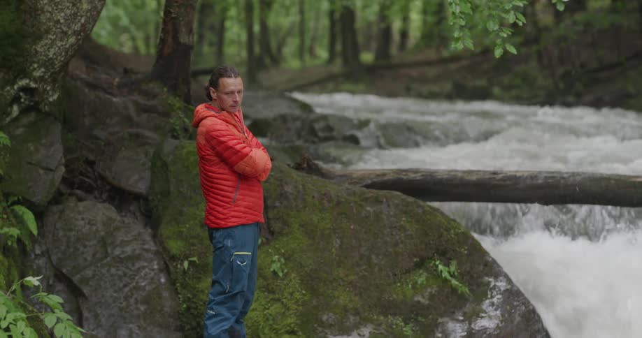 A man wearing a striped orange shirt and jeans stands on the side of a river and big boulders