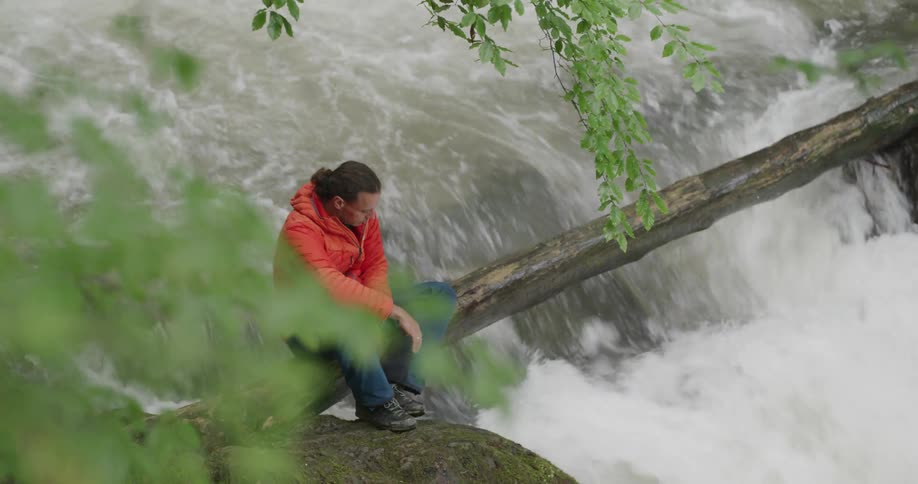 A man sitting on a log spanning a waterfall sticks his hand into the water running beneath him
