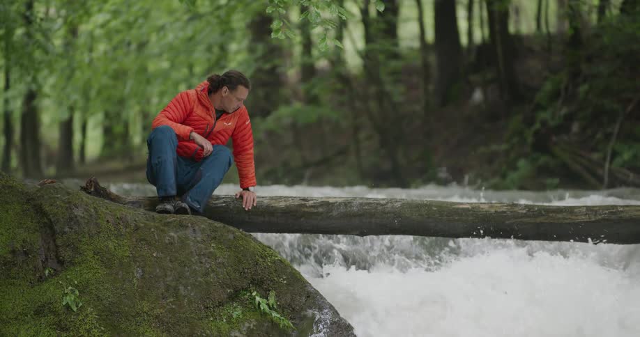A man sits on a fallen tree that is located over a rapidly rushing river