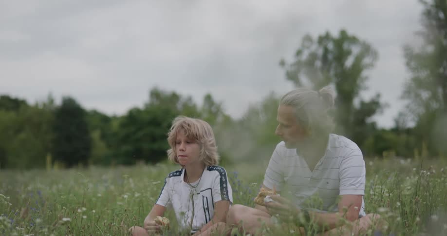 Father and son sit in grass eating sandwiches father reaches over to steal a piece of the sons sandwich and the son pulls it away making them both laugh