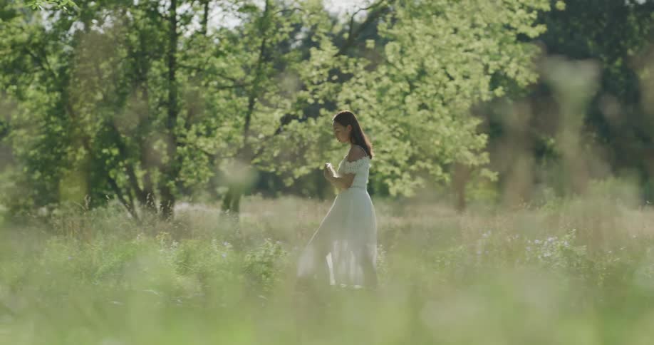 Female person wearing long white dress walks through field surrounded by trees and woods and sits down in the grass