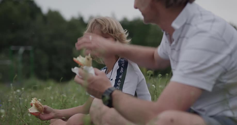 Father and son sit in grass and eat sandwiches after toasting them with trees in the background