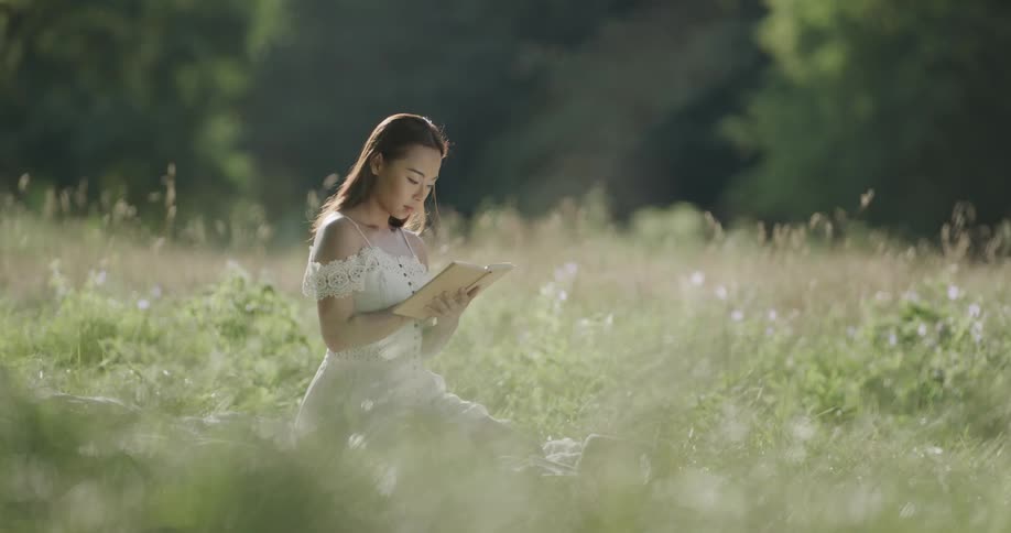 A woman sitting in a grassy field reading a book with trees in the background