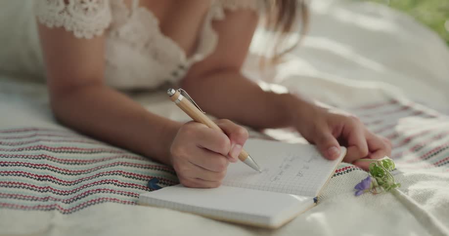 A woman wearing a white dress lies on a blanket outdoors writing in a notebook during the day