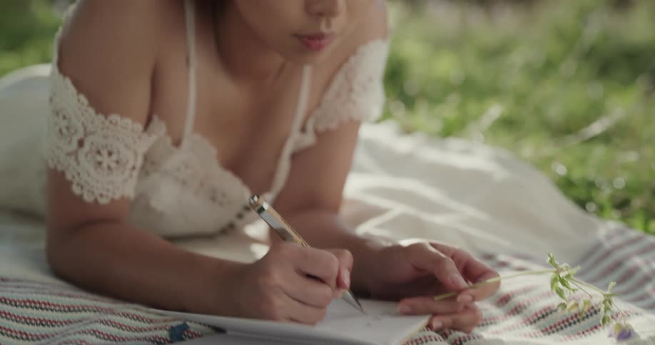 Woman lays on blanket writing in notebook in a grassy area on a sunny day