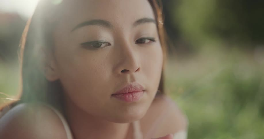 A female asian in a white dress laying on the grass twirling a flower