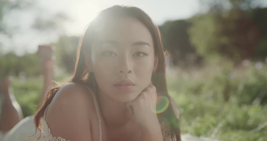 An asian woman laying on her stomach on a blanket in the grass surrounded by tall grass and trees