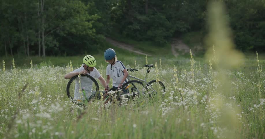 Father and son fix a bicycle while outdoors in a field as they wear helmets