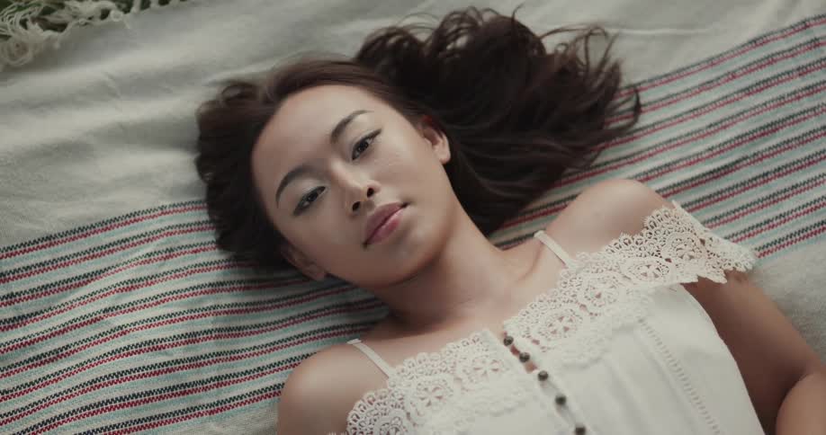 A young asian woman rests with her eyes closed on a blanket laid in a field before slowly opening her eyes and looking into the camera