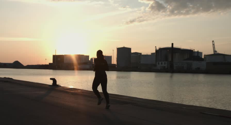 A woman jogs along a river boardwalk in an industrial area at dawn