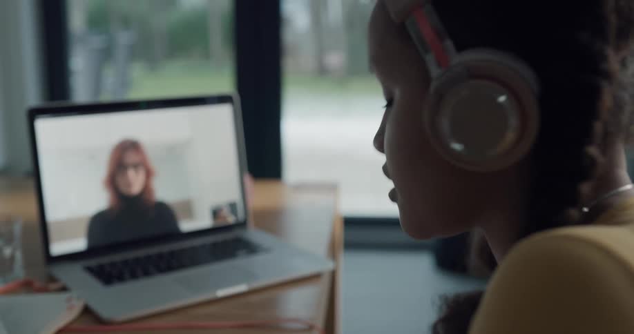 A girl writes in a workbook while using a laptop to speak to a woman through video conference