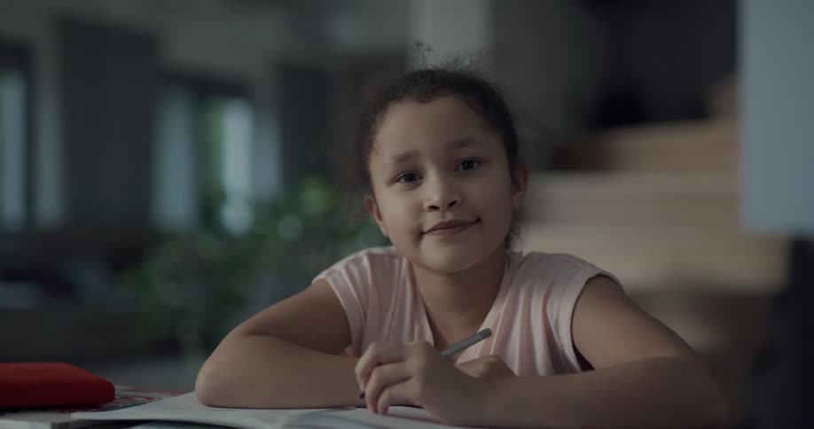Young girl doing homework on a table in a room at home looks at the camera and smiles