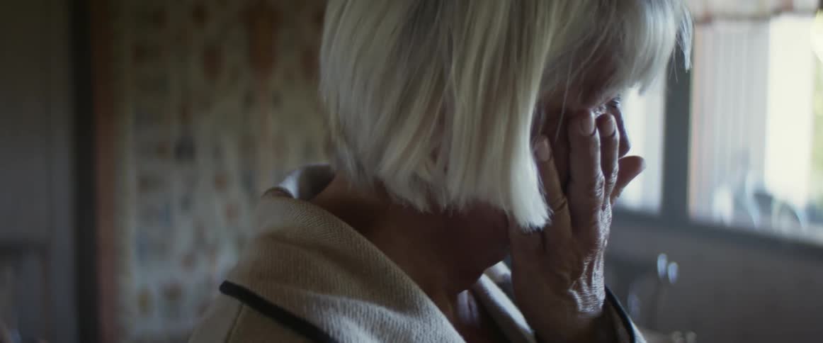 An elderly woman stands in her kitchen and cries