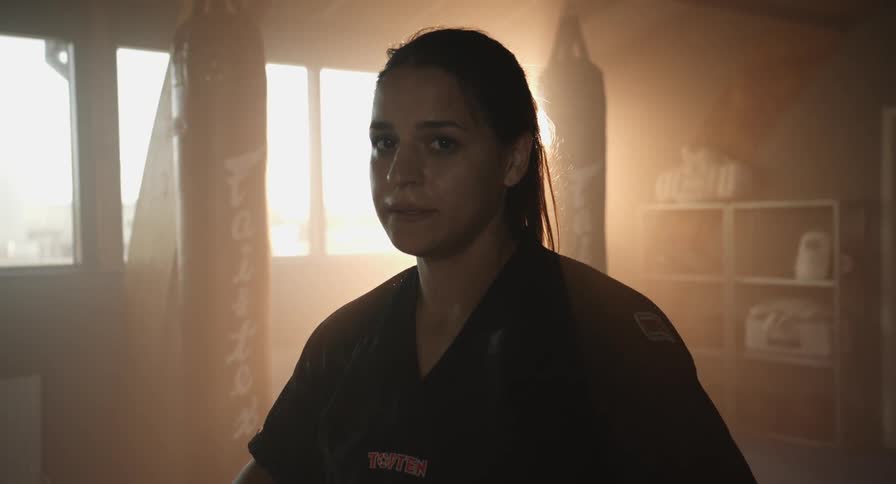 A female boxer stares into the camera and breathes heavily while standing in a gym