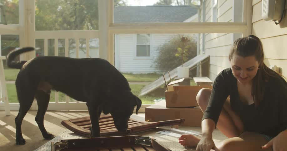 A young woman sitting on the enclosed porch of a house pets a dog before preparing to cut open a box with a pair of scissors