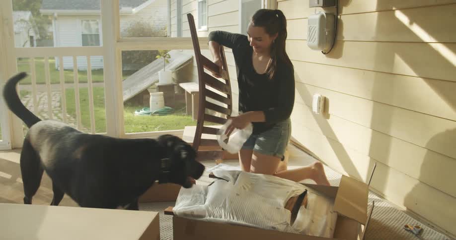 A woman laughs as her large black dog walks around her as she removes parts of a chair from a box while sitting in a screened in porch
