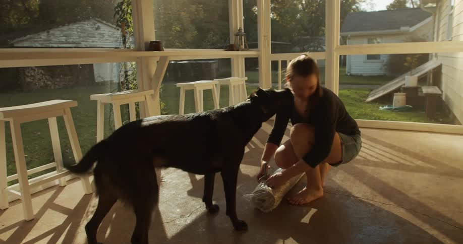 A woman on a porch with a dog opening a carpet in plastic with scissors