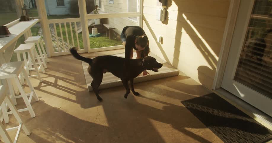 A woman unrolls a large rug on the floor of a patio while her dog looks on
