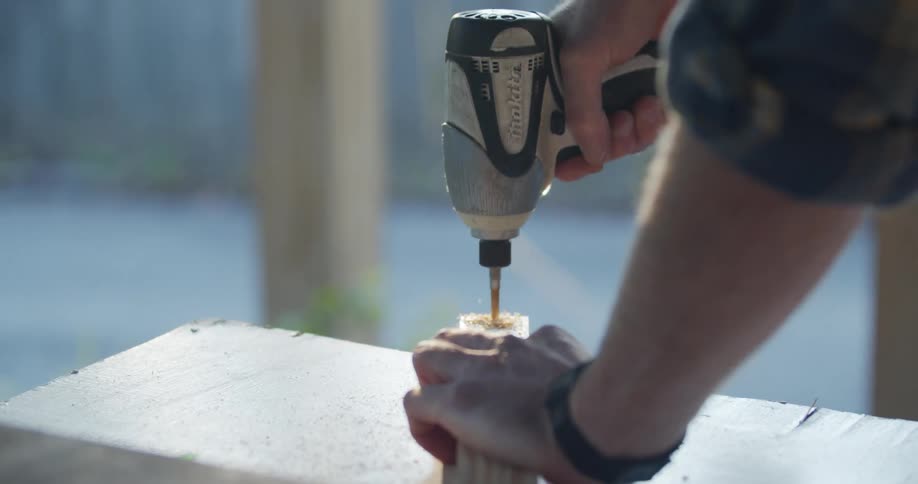 A man uses a power drill to drill a hole in a piece of wood outside