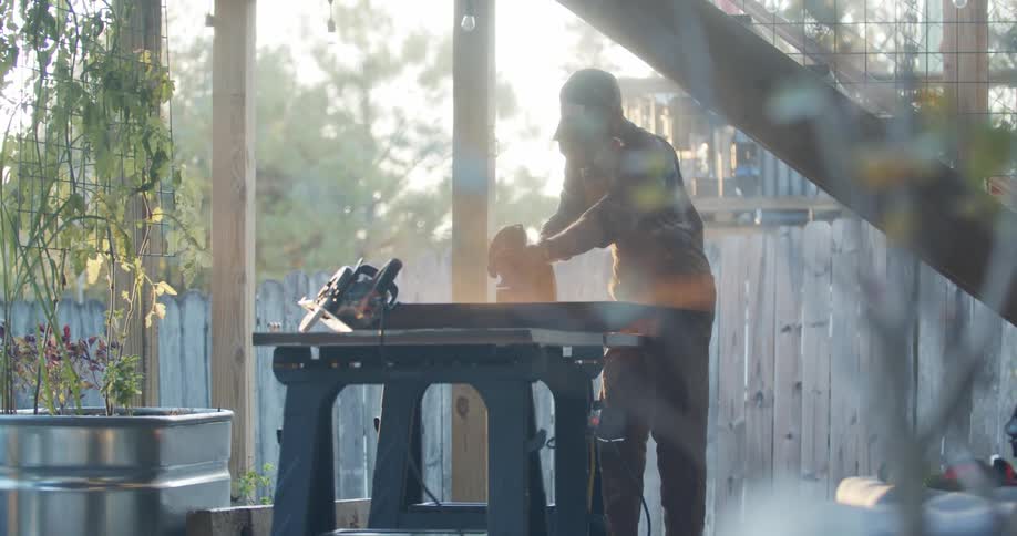 Man with a hat sanding an object on a table while outside in the morning sun