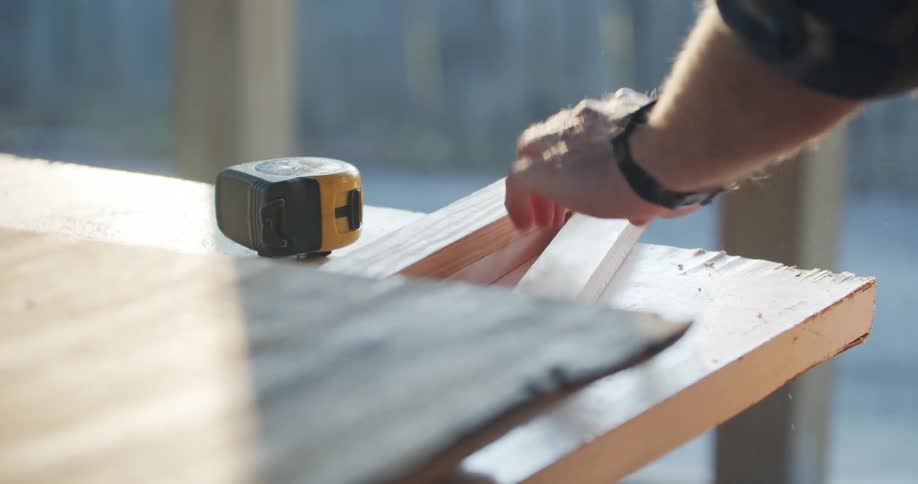 Carpenter uses a wireless drill to drill a hole in wooden boards outdoors at sunset