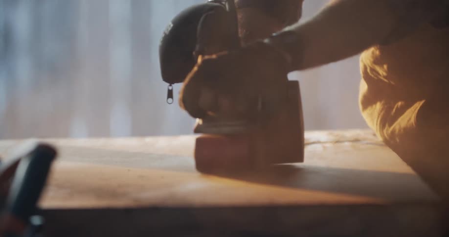 Man wearing gloves uses an electric sander on a piece of plywood