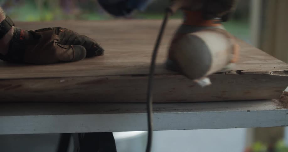 A man with gloves a hat and a mask sanding a table with an electric sander