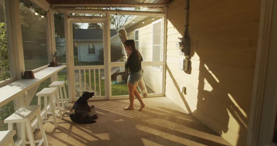 A young woman carries a rolled up rug out to her sunroom where her dog is lying