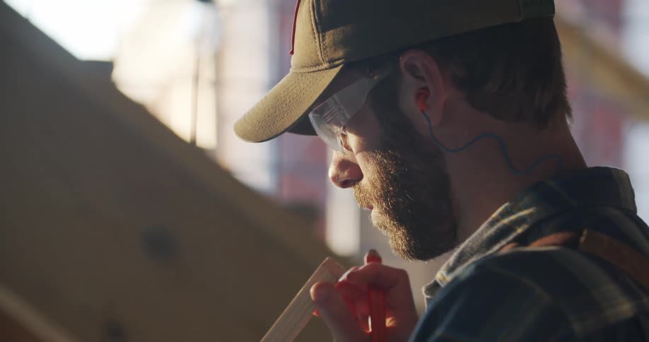A man holds a piece of wood and a marker while wearing safety glasses and a hat at a construction site outdoors