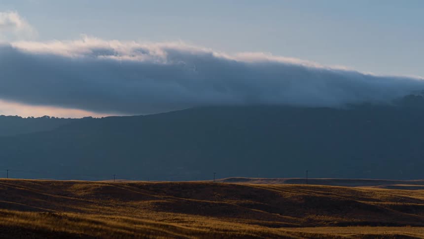 Clouds move across the top of a mountain top in the distance from a field of small hills and brown grass