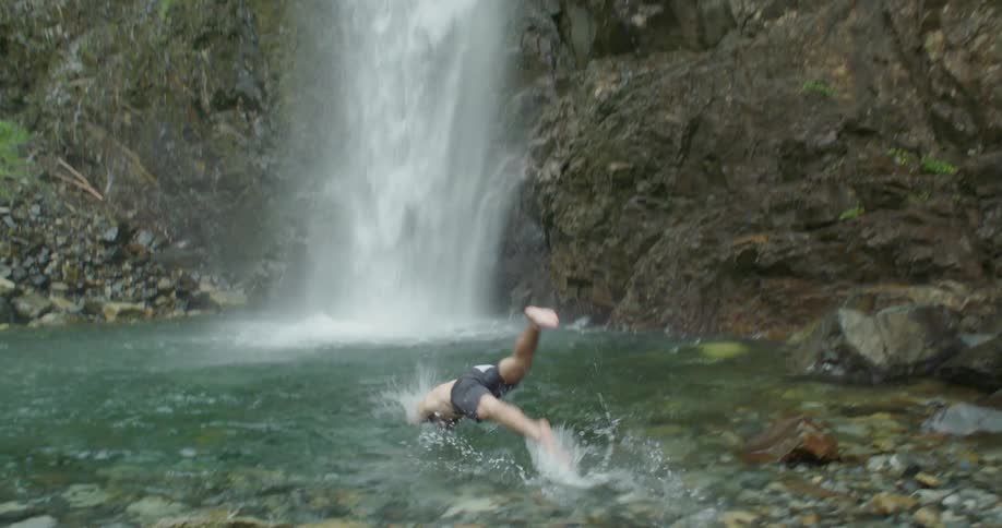 A man jumping into water right by a waterfall