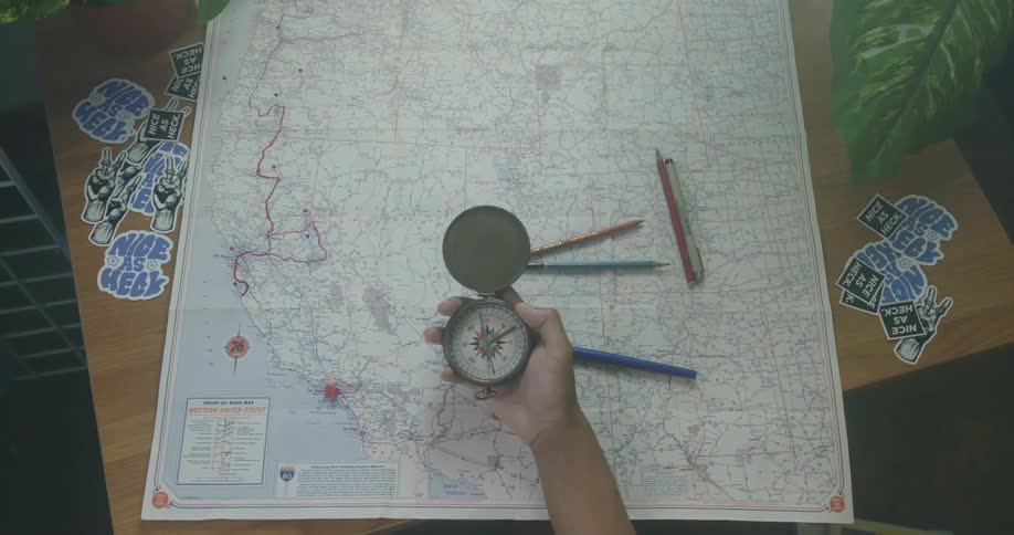 A person holds a compass in their hand over a map and pens lying on a table
