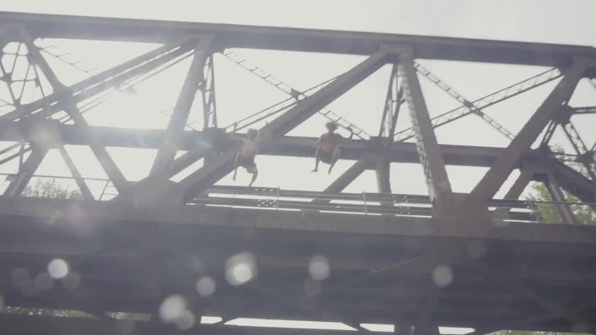 Two boys in swim trunks jump from a tall bridge into a murky river