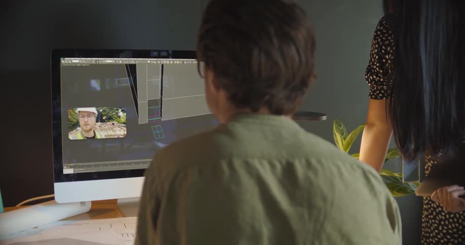 A man and work work in an office looking at building schematics with another worker via laptop