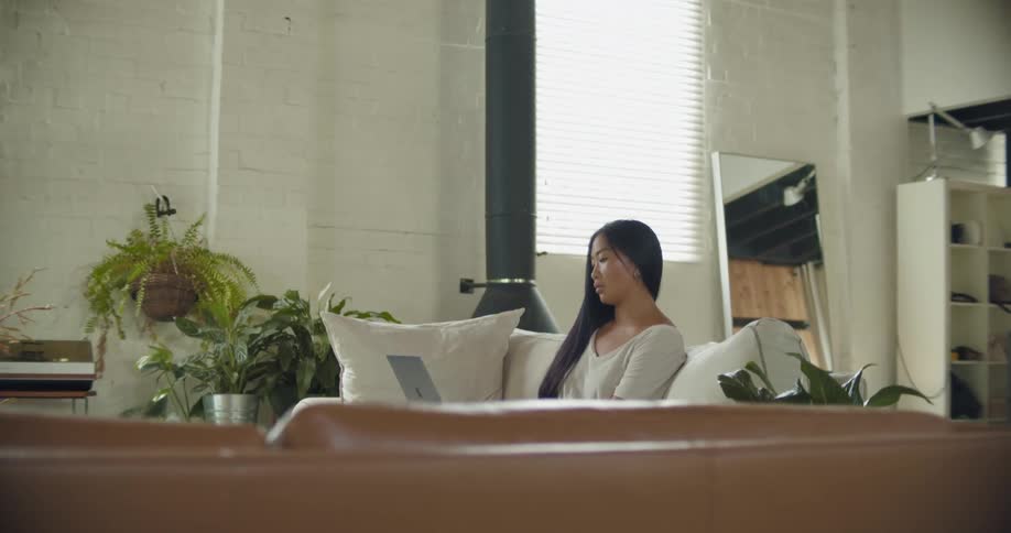 A young woman using a laptop while sitting on a sofa in the living room of a house stops to check her phone