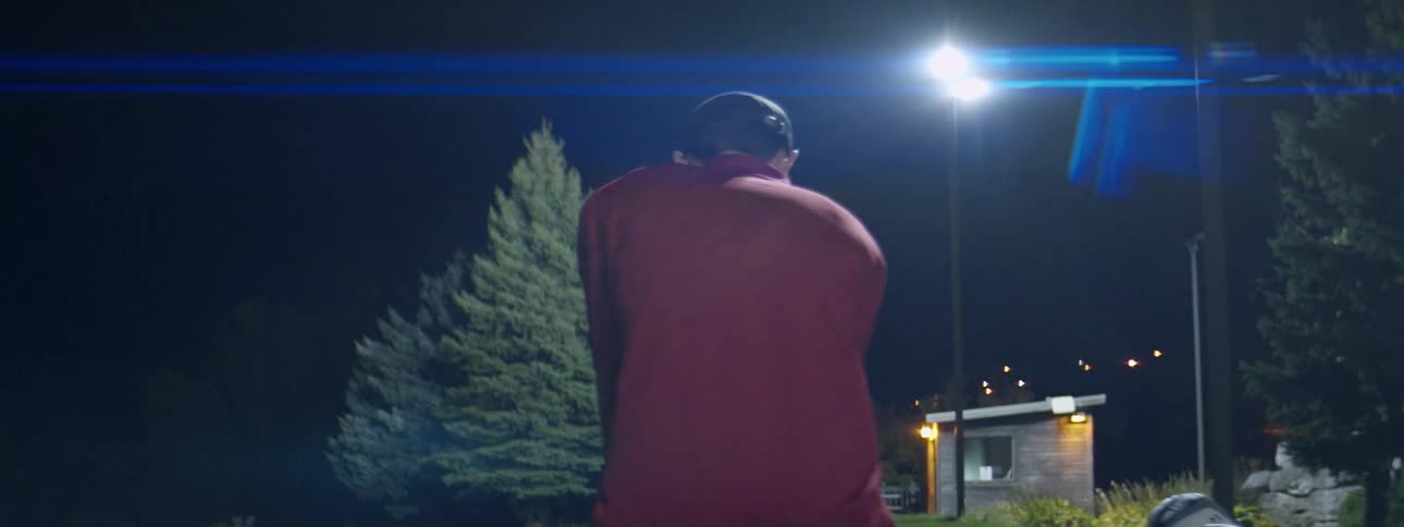 A man wearing a red jacket and a baseball hat swings a gold club at a driving range at night
