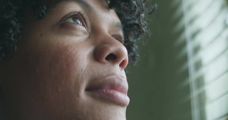 A teenager with short curly hair sits next to window blinds and looks out and up