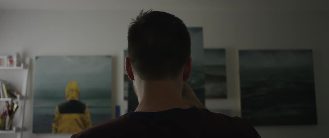 A man stands in front of a wall full of photos looking at them and drinking from a cup