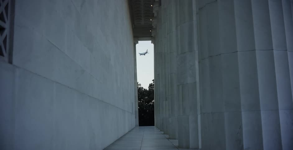 A small plane flies past a large building with ridged columns