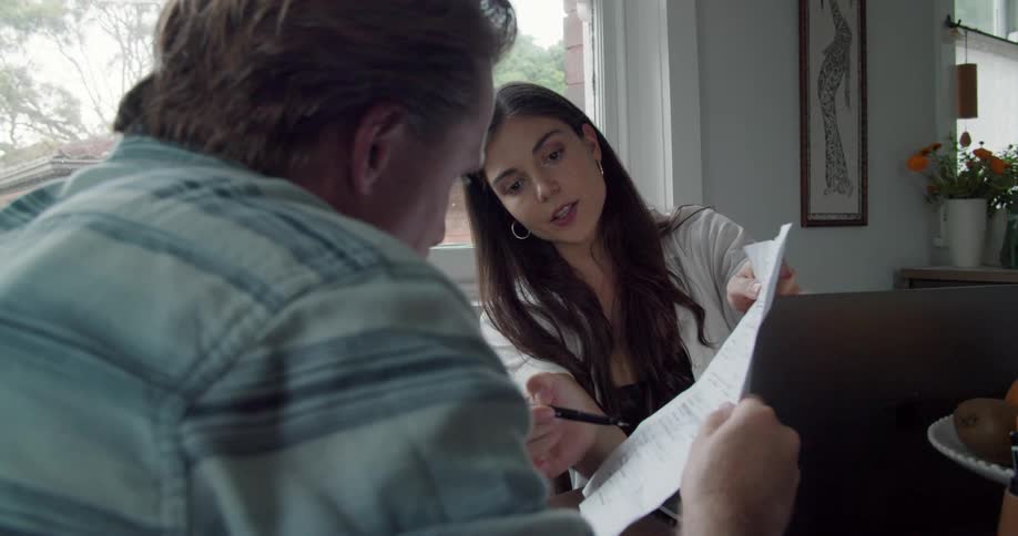 A woman shows and explains a paper form to a man as they sit together at a table indoors during the day