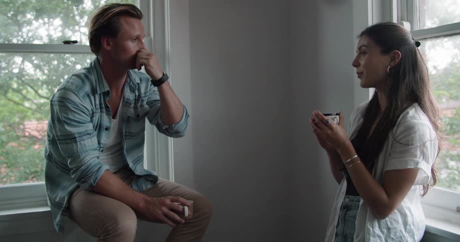 A man and woman chat while drinking coffee next to windows indoors