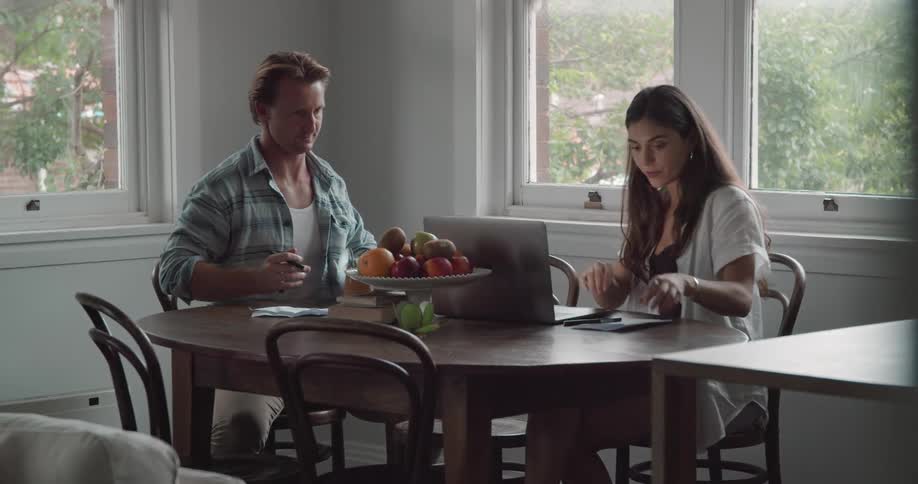 A man and woman sit at a table at home and the woman looks at a laptop and the man hands her a book and leans over and looks at the computer