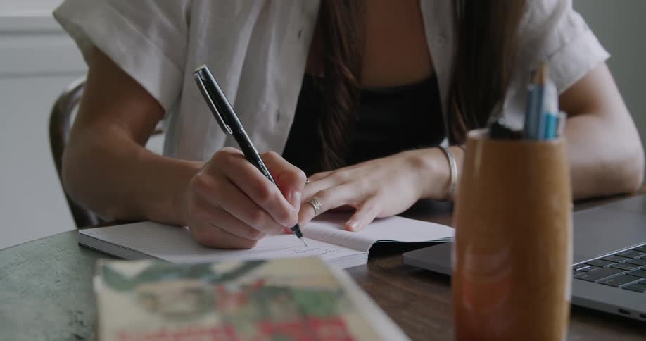 A woman takes a sip from a drinking glass before writing in a notebook while glancing at the screen of a laptop as she sits at a wooden table in a house during the day