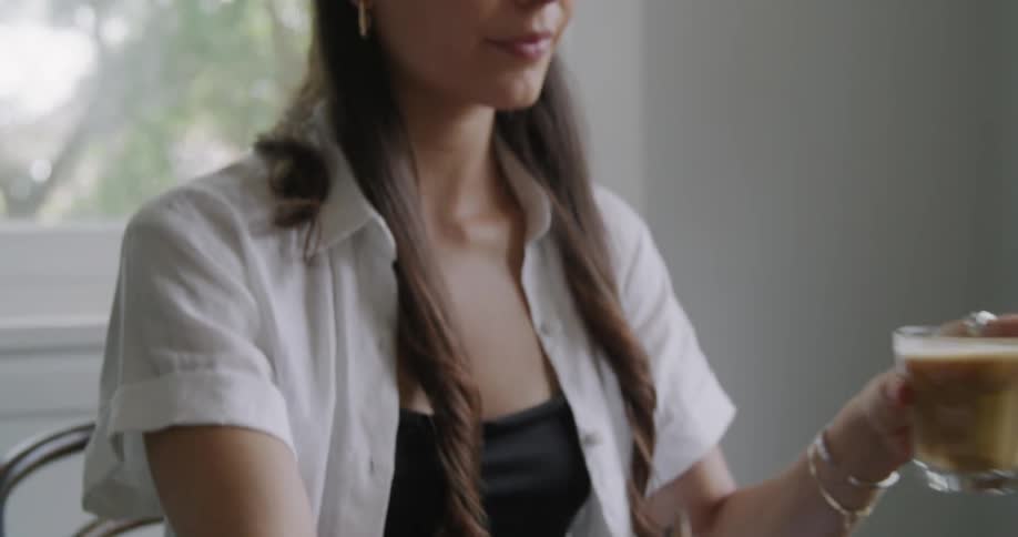 A woman sits at a table typing on a laptop and drinking coffee
