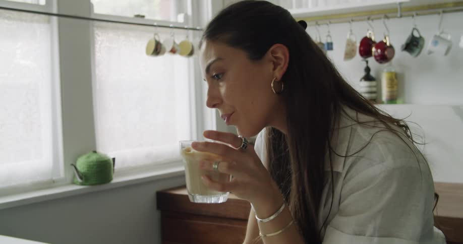 Brunette woman with long hair and white shirt uses a laptop while drinking beverage in kitchen