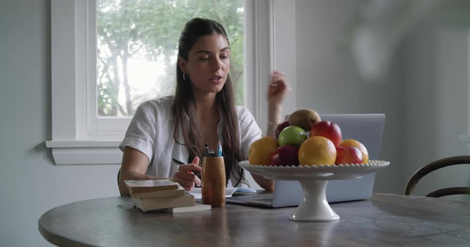 A woman sits at a table taking notes while communicating on a laptop